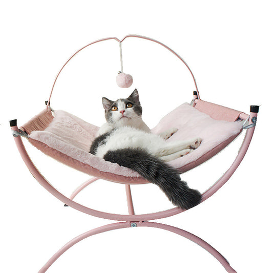 A calm cat resting on a pink, fluffy cat bed, gazing thoughtfully with a dangling pom-pom nearby.