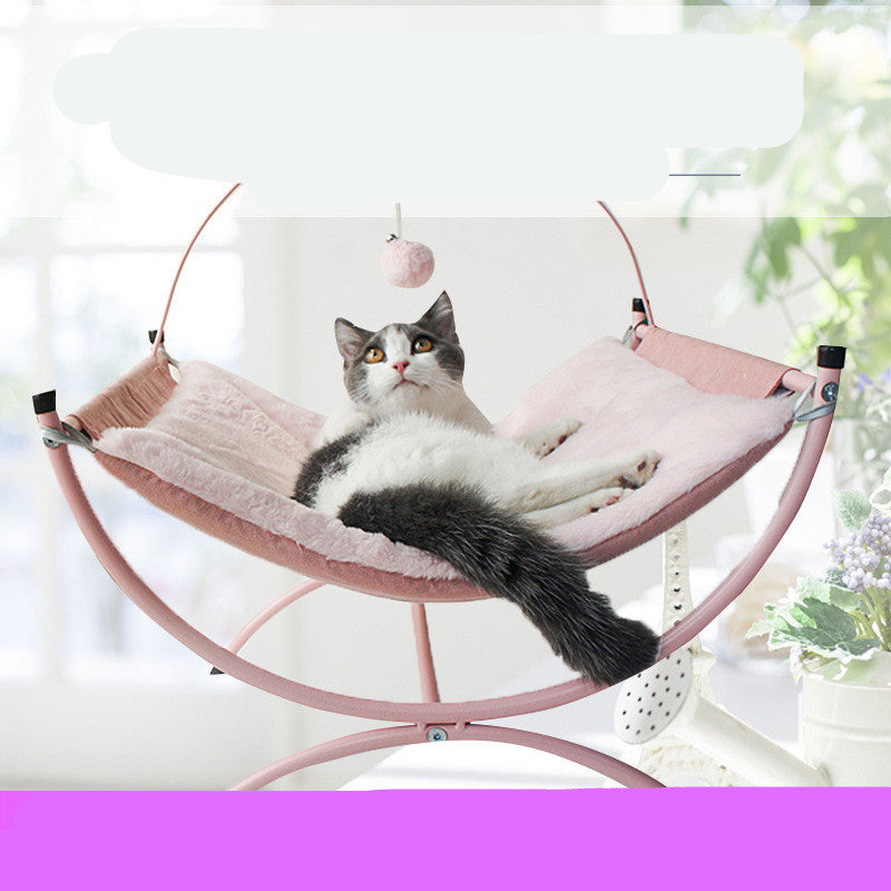 A calm cat resting on a pink, fluffy cat bed, gazing thoughtfully with a dangling pom-pom nearby.