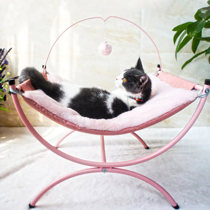 A calm cat resting on a pink, fluffy cat bed, gazing thoughtfully with a dangling pom-pom nearby.