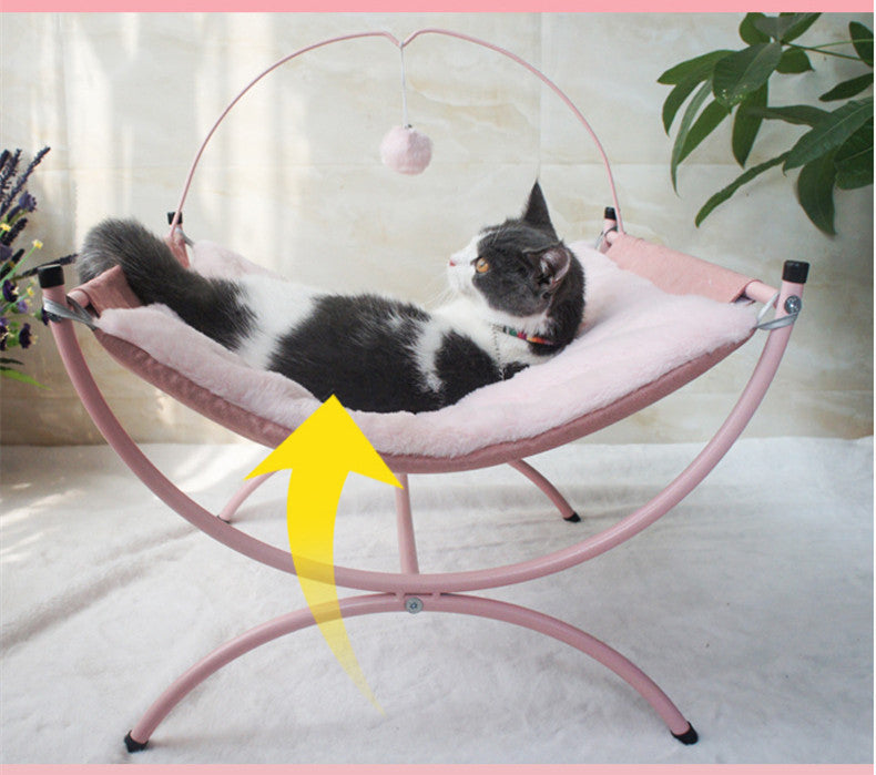 A calm cat resting on a pink, fluffy cat bed, gazing thoughtfully with a dangling pom-pom nearby.